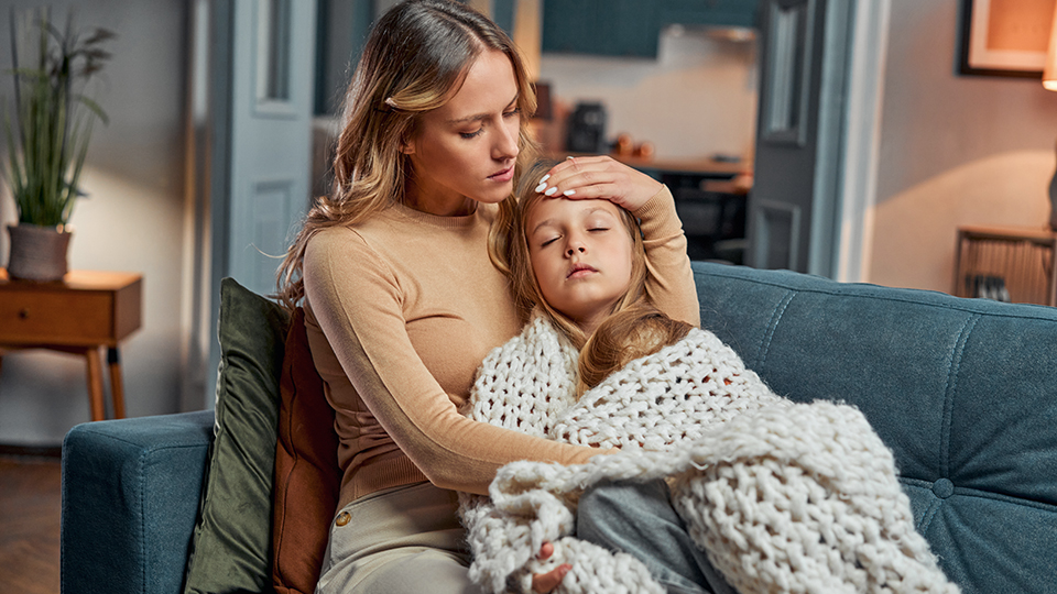 Mother comforts her sick child resting on a couch with a blanket at home