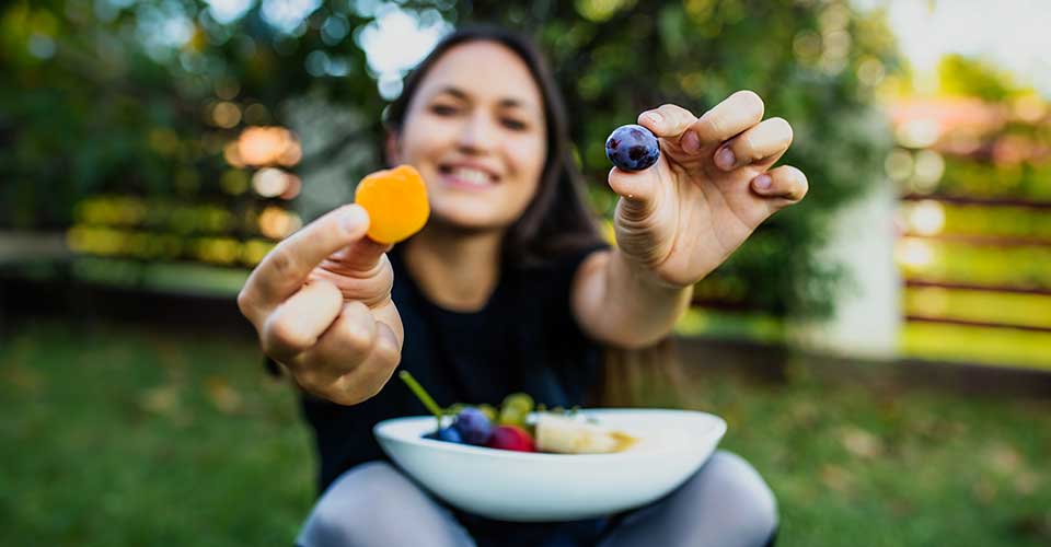 Teen girl with bowl of fruit in lap, holding up a blueberry and an apricot