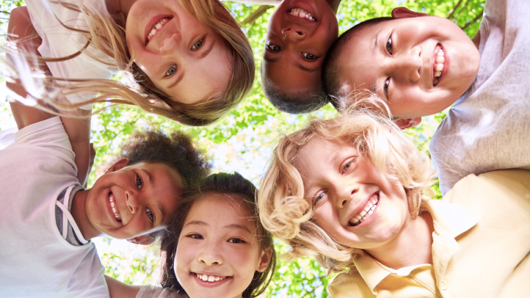 Group of diverse, smiling children looking down at the camera under a tree – promoting healthy kids' smiles for pediatric dental care in Fleming Island, FL