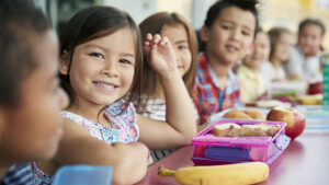 Smiling elementary school children sitting at a lunch table with healthy lunchboxes filled with fruit, sandwiches, and balanced snacks.