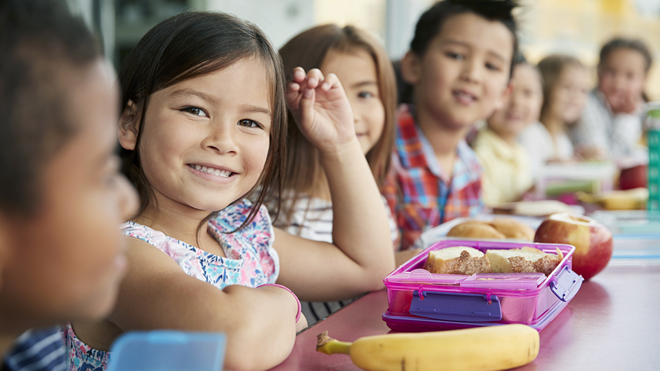 Smiling elementary school children sitting at a lunch table with healthy lunchboxes filled with fruit, sandwiches, and balanced snacks.